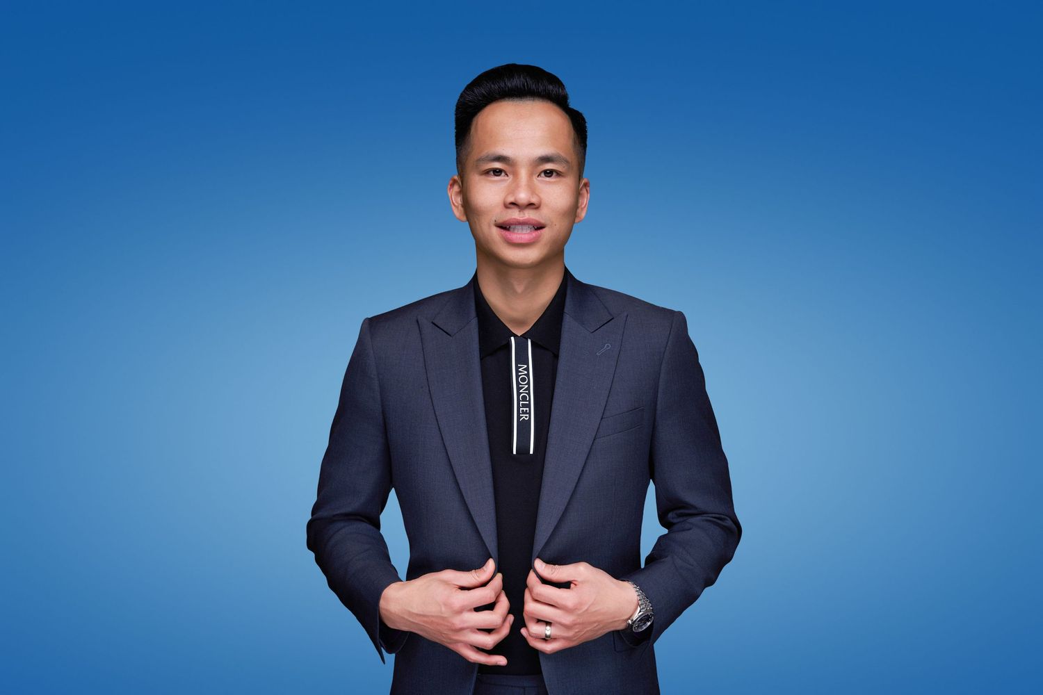 A person in a navy suit and black shirt poses confidently against a blue background.