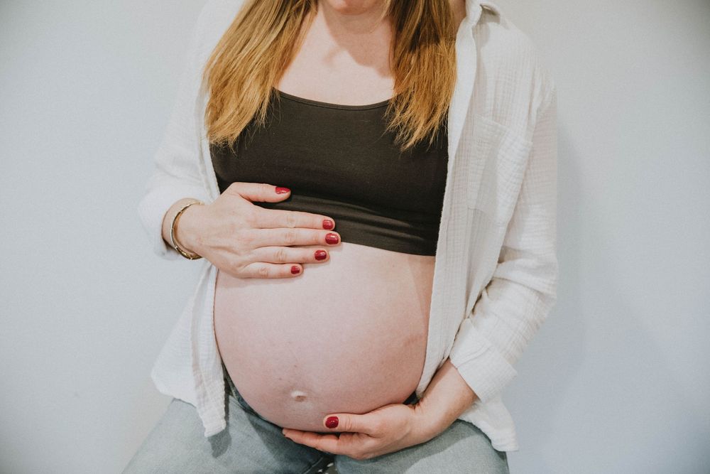 An expectant parent in a white cardigan and gray pants lovingly cradles their pregnant belly while looking down. indiana pa photographer