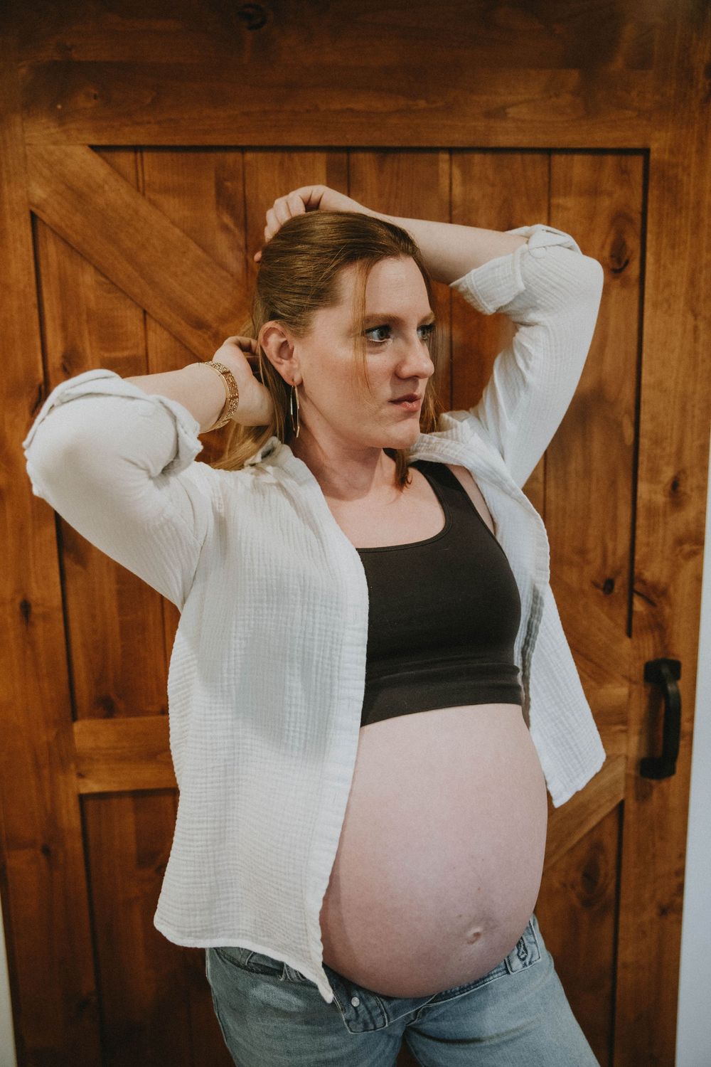 A pregnant person in a white cardigan and black crop top poses against a rustic wooden barn door. indiana pa photographer, maternity session