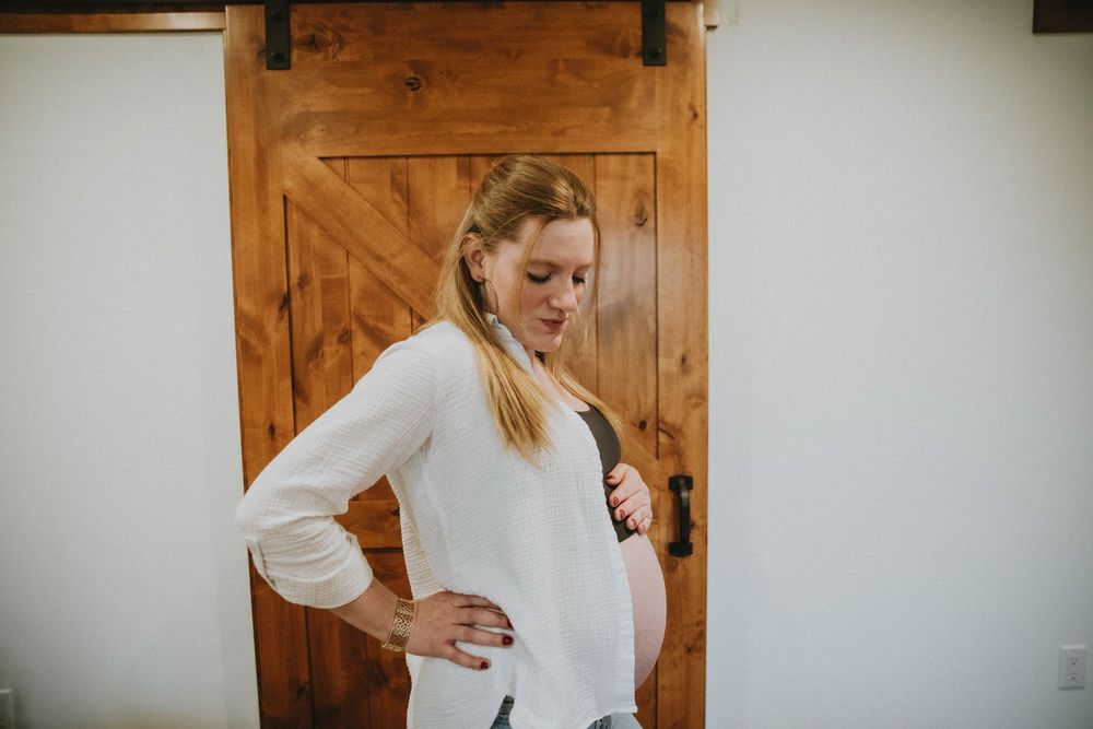 A pregnant person in a white cardigan and black crop top poses against a rustic wooden barn door. indiana pa photographer, maternity session