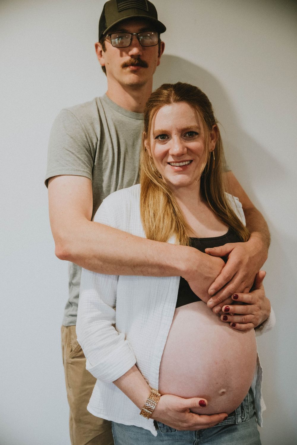 A pregnant person in a white cardigan and black crop top poses against a rustic wooden barn door. indiana pa photographer, maternity session