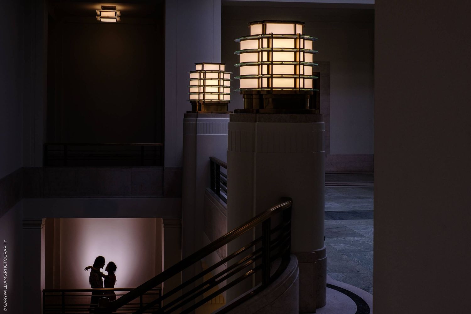 Two silhouettes walk past a hallway with illuminated geometric windows casting dramatic lighting patterns.