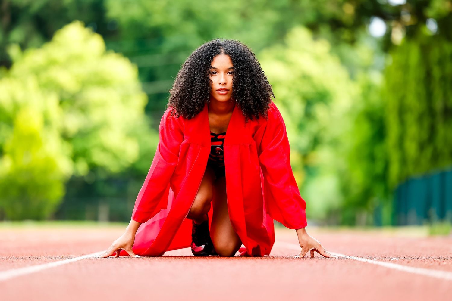 Person in a bright red suit jacket poses dramatically on a running track in a lush green outdoor setting.