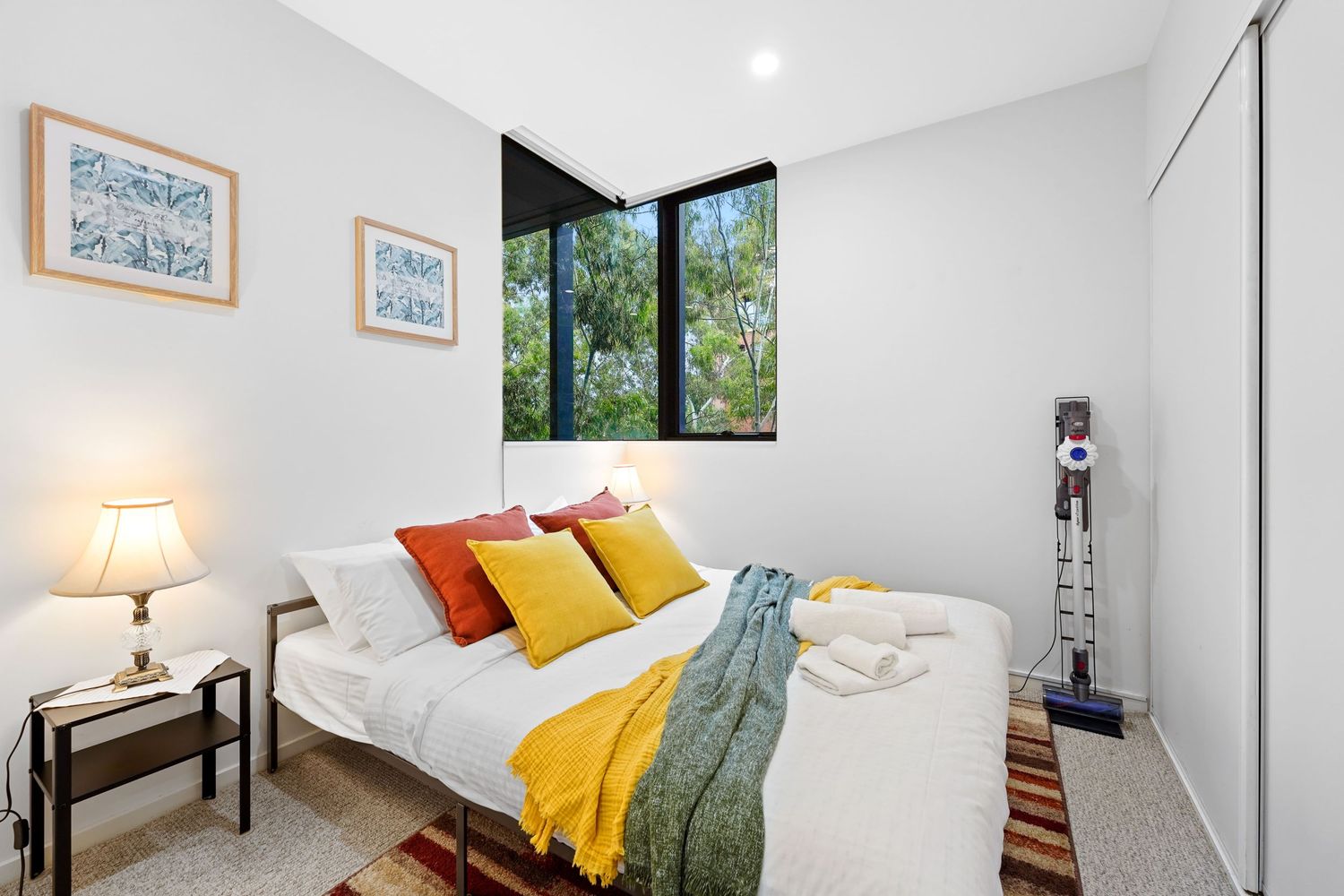 Modern bedroom with white walls, corner windows, and colorful accent pillows in yellow and orange on white bedding.