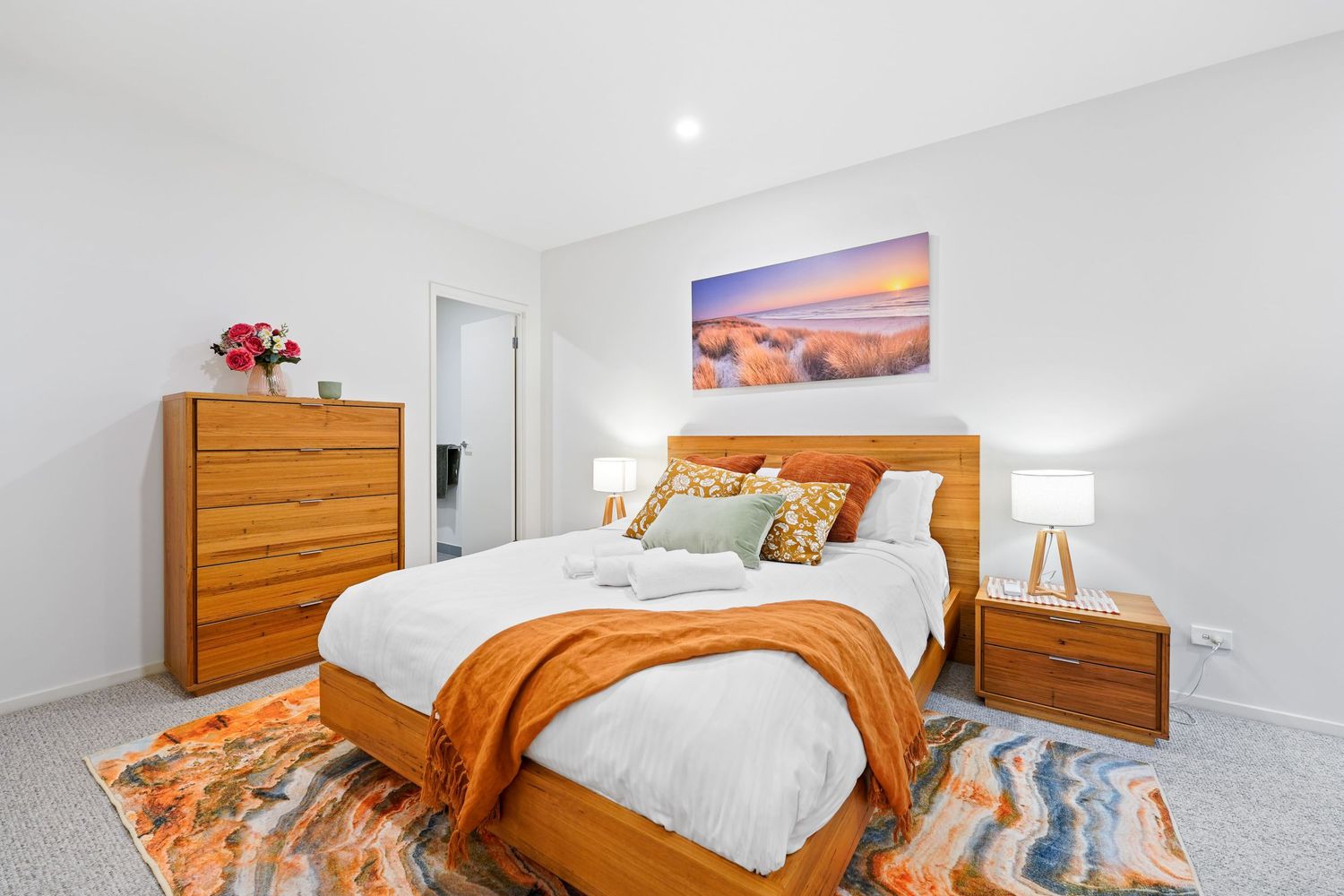 Modern bedroom with wooden furniture, orange accents, and coastal artwork featuring a sunset beach scene above the bed.