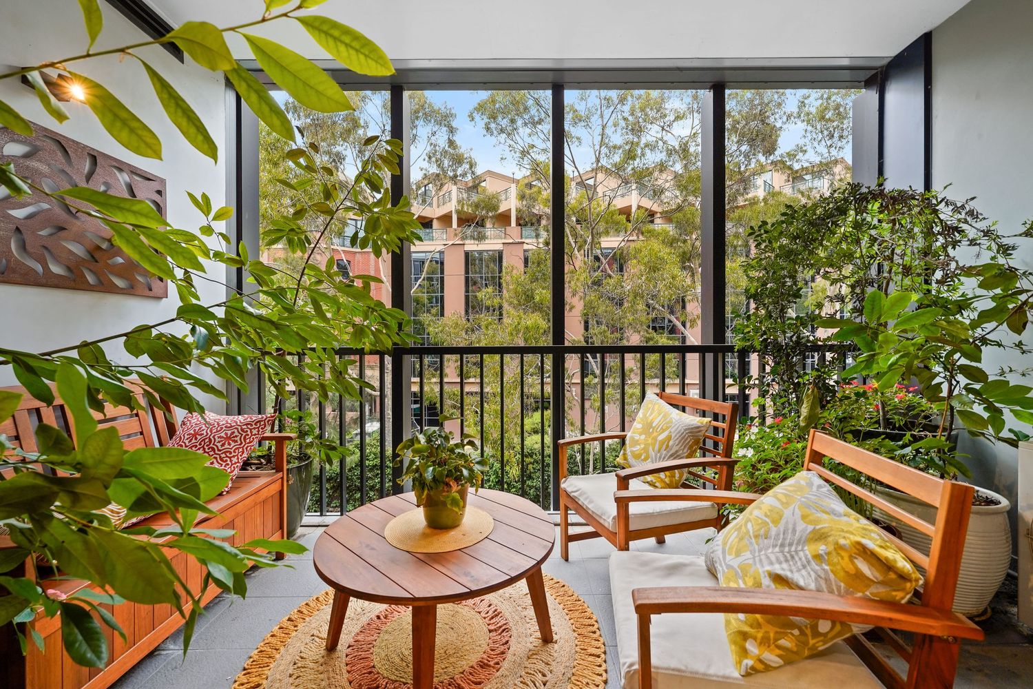 Cozy screened balcony with wooden furniture, green plants and scenic view of trees and buildings in the background.
