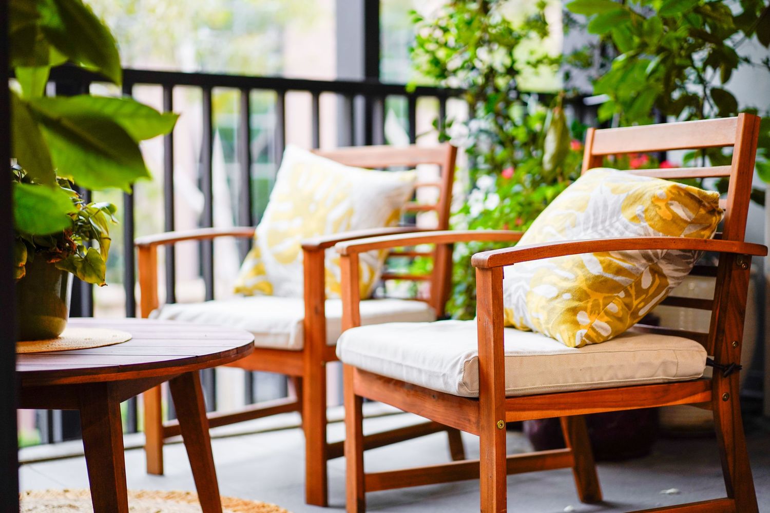 Wooden patio chairs with white cushions and decorative pillows on a cozy balcony with green plants.