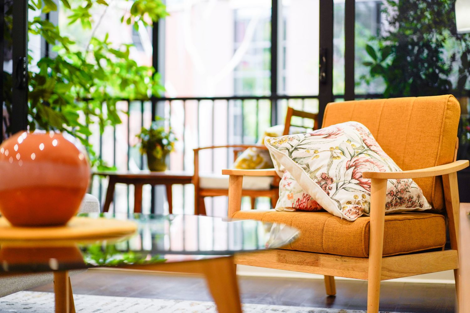 Mid-century modern orange armchair with decorative pillow on a balcony with green plants and wood side table.