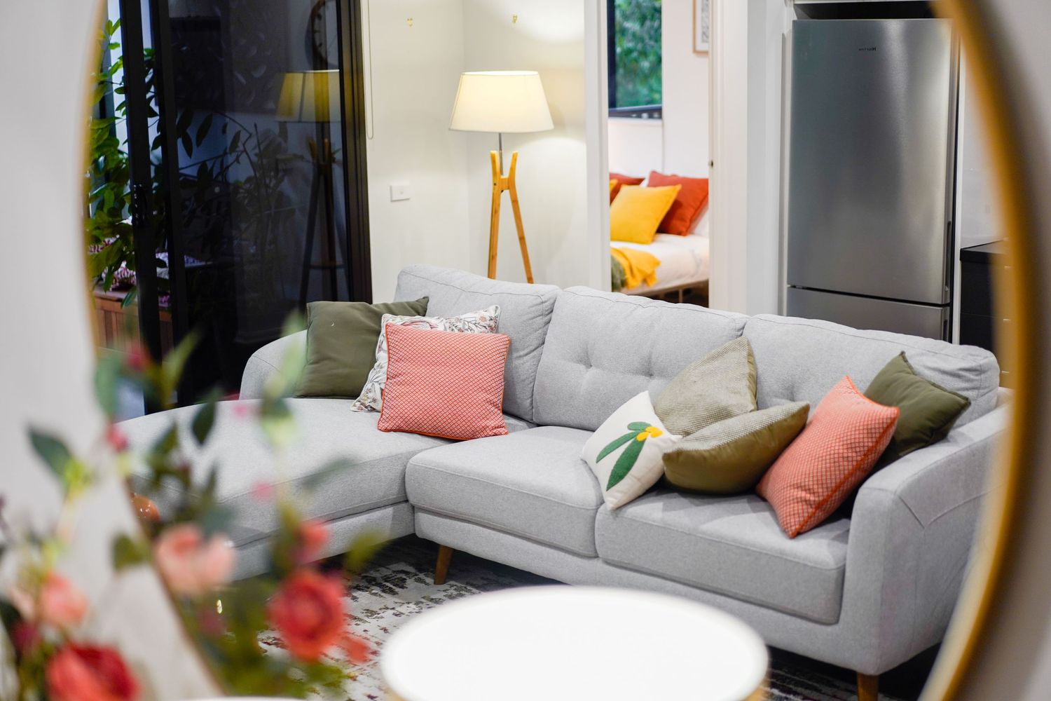 Modern living room interior with gray sectional sofa, coral accent pillows, and floor lamp reflected in round mirror.