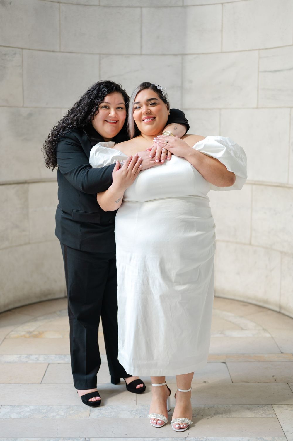 Two women embrace while posing against a curved stone wall, one in a white dress and one in black attire during engagement session