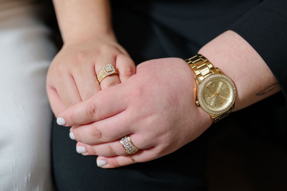 Close-up of folded hands showing gold rings and a gold watch against dark and light clothing during engagement session