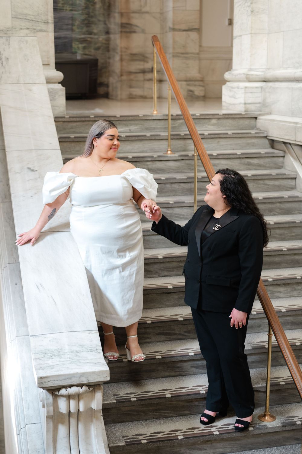 Two people stand on classical stone steps, one wearing a white dress and one in black, sharing a joyful moment together at NY Public Library