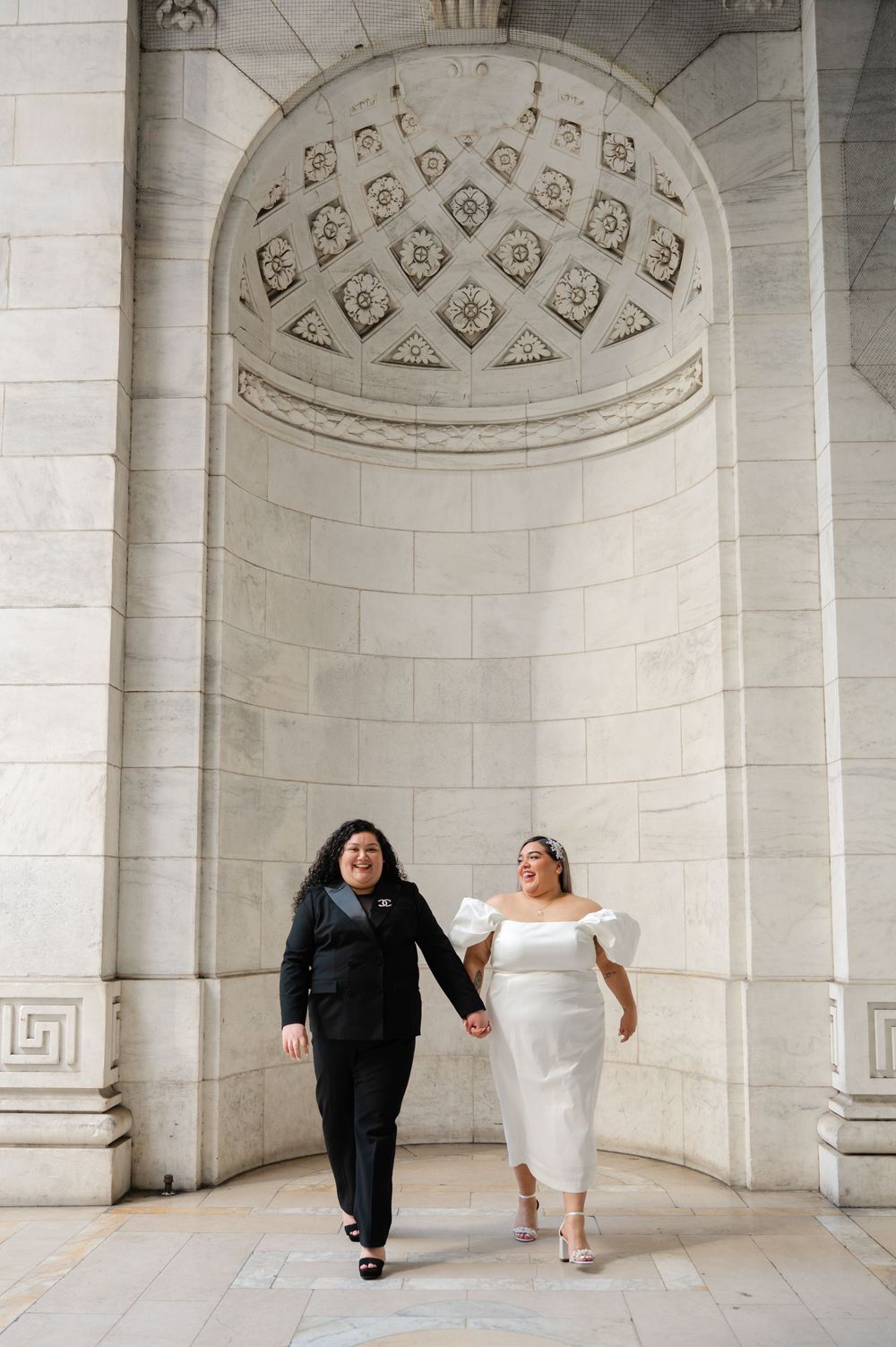 Two happy newlyweds hold hands and walk together under an ornate archway with decorative ceiling details at NY Public Library