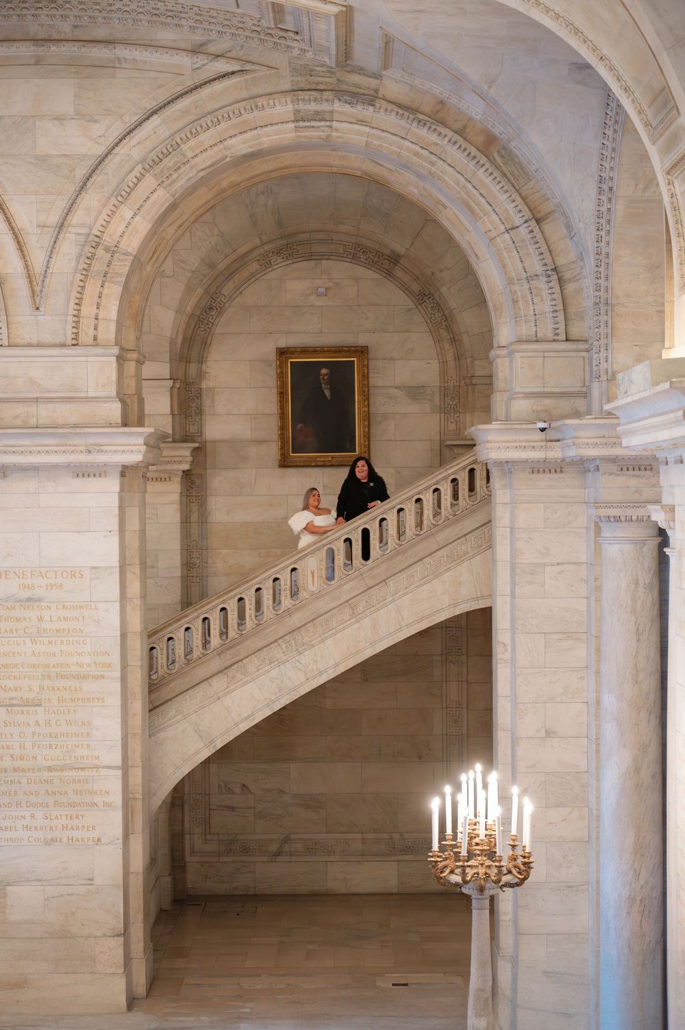 Couple in an elegant stone interior with arched ceilings, ornate staircase, and chandelier at New York Public Library in engagement photos