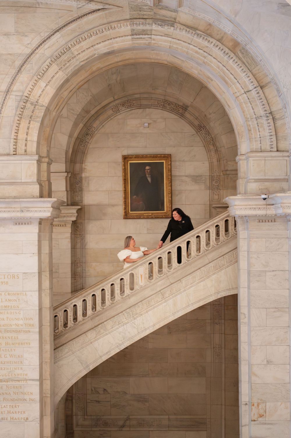 Two people pose on an elegant curved staircase under an arched ceiling with a portrait hanging on the wall at NY Public Library