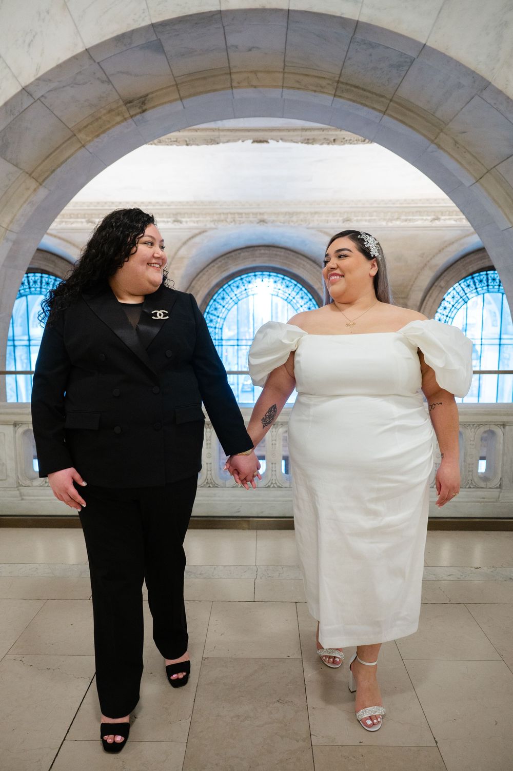 Two people in contrasting black and white outfits hold hands while standing under elegant stone arches with blue windows at NYPL.