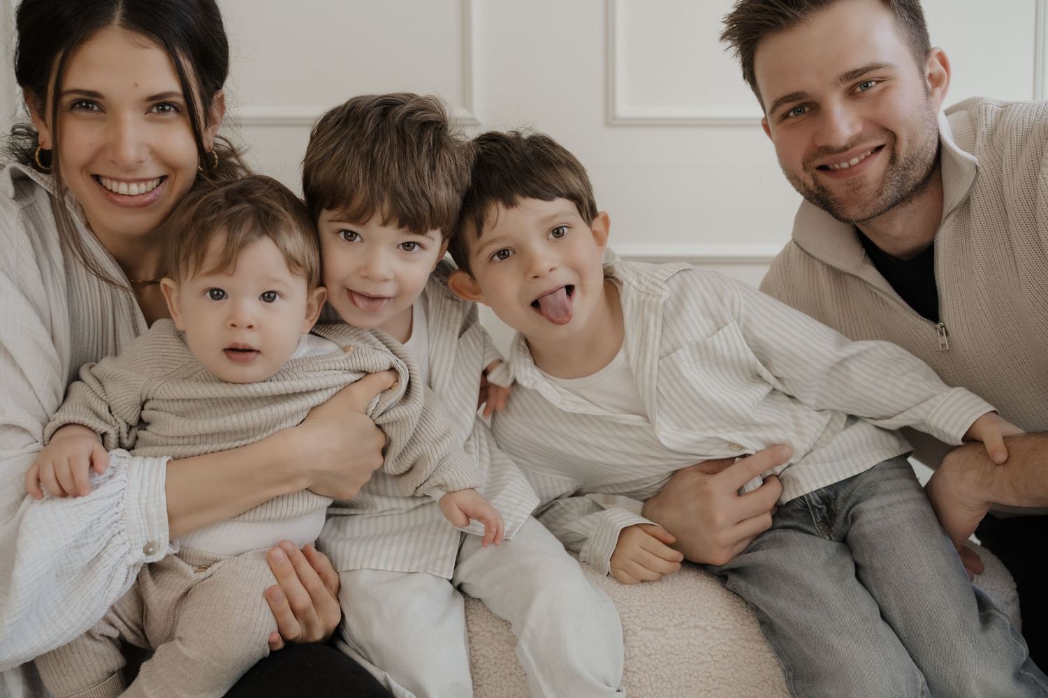 A happy family of five wearing matching light colored casual outfits sitting close together at home.