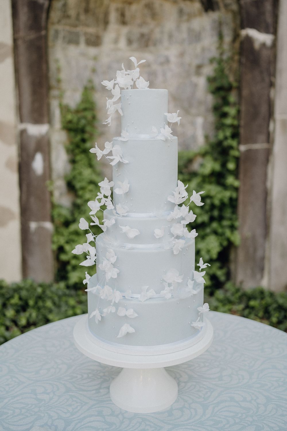 A tall, elegant, five-tiered pale blue wedding cake adorned with delicate white sugar flowers cascading down its sides,