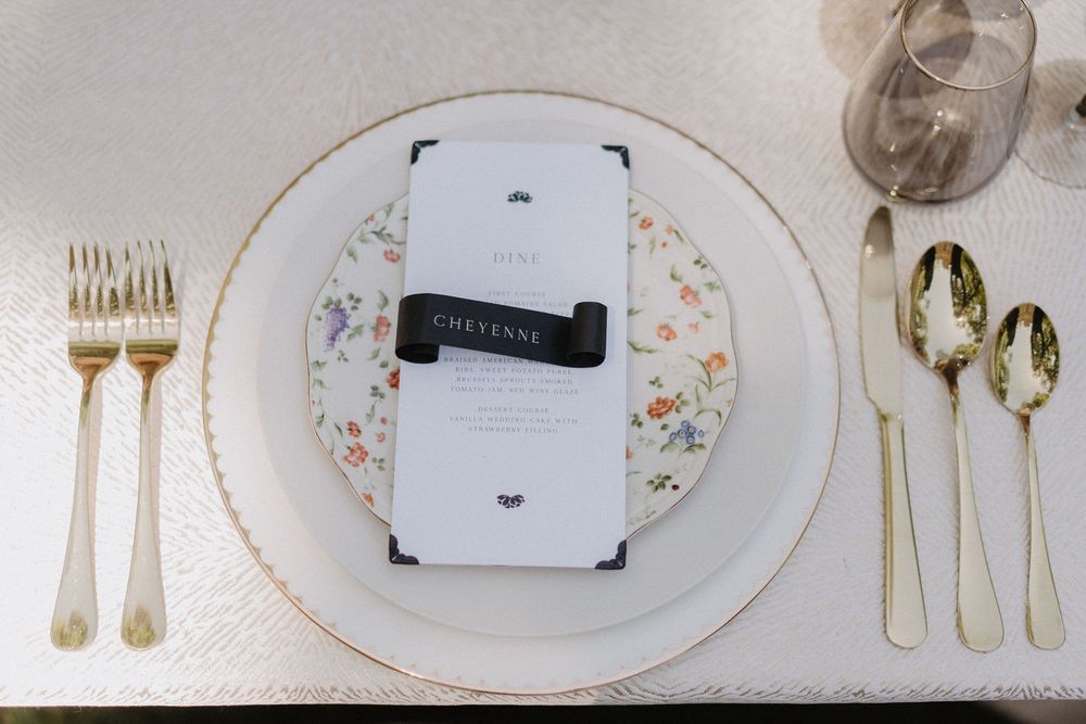 A formal place setting with gold flatware and a floral china plate featuring a menu card.