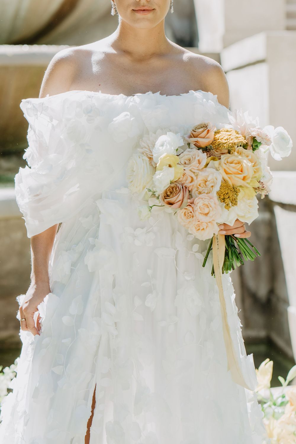 A bride in an off-shoulder white wedding dress decorated with floral appliqués, holding a romantic bouquet of soft peach