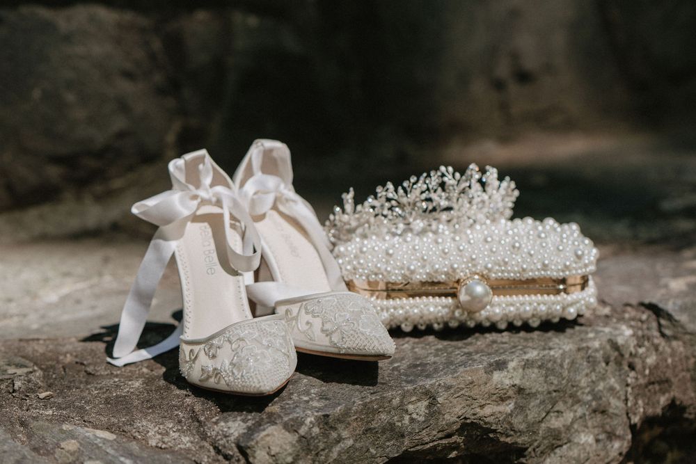 A pair of ivory lace wedding shoes with ankle ribbon ties displayed alongside a pearl-encrusted clutch bag