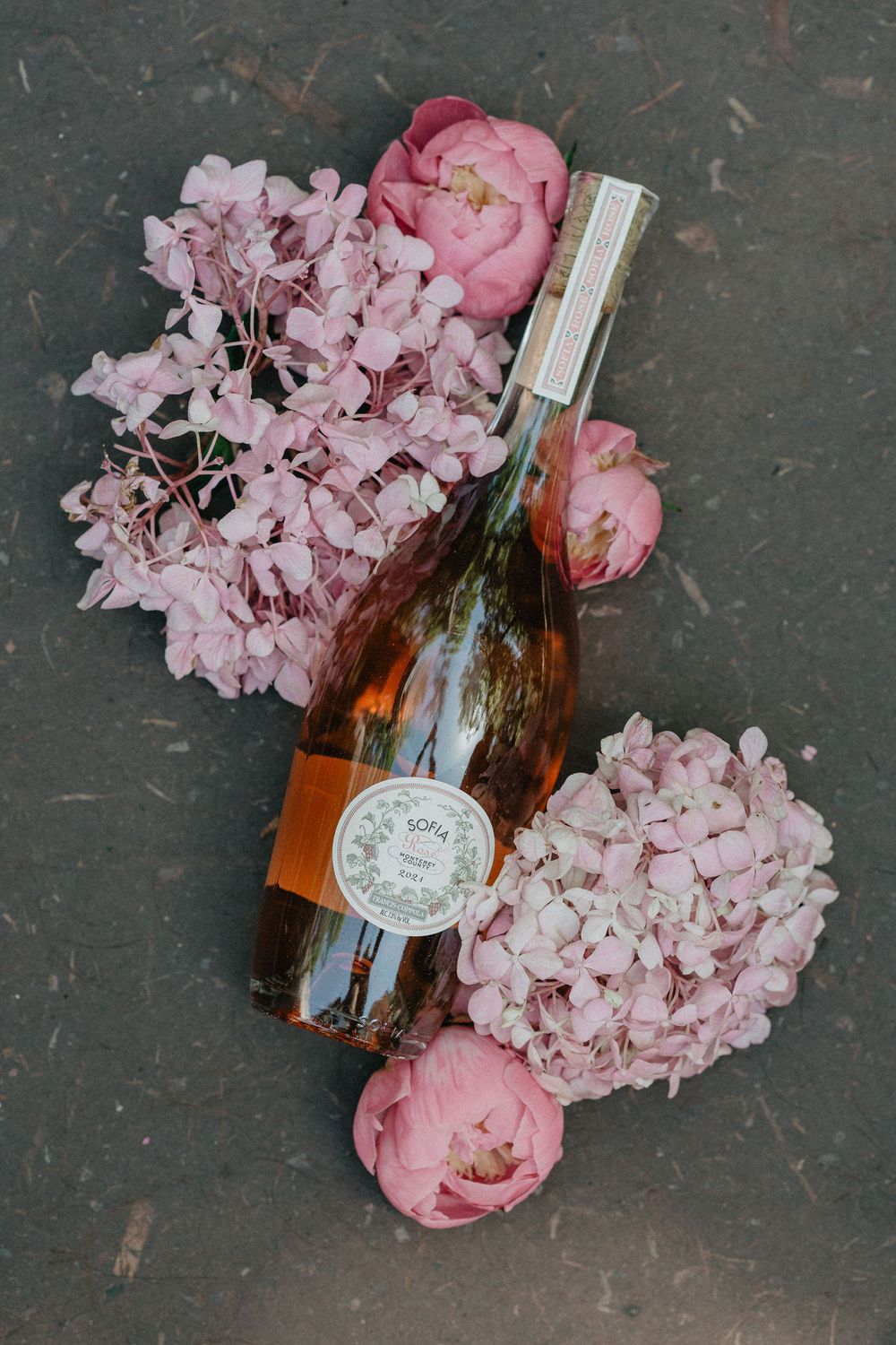 Pink hydrangeas and peonies arranged with a wine bottle on dark surface.