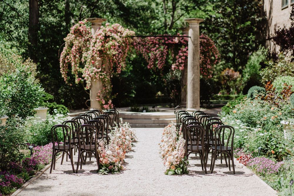An outdoor wedding ceremony setup with rows of black chairs and floral arch in a garden setting.
