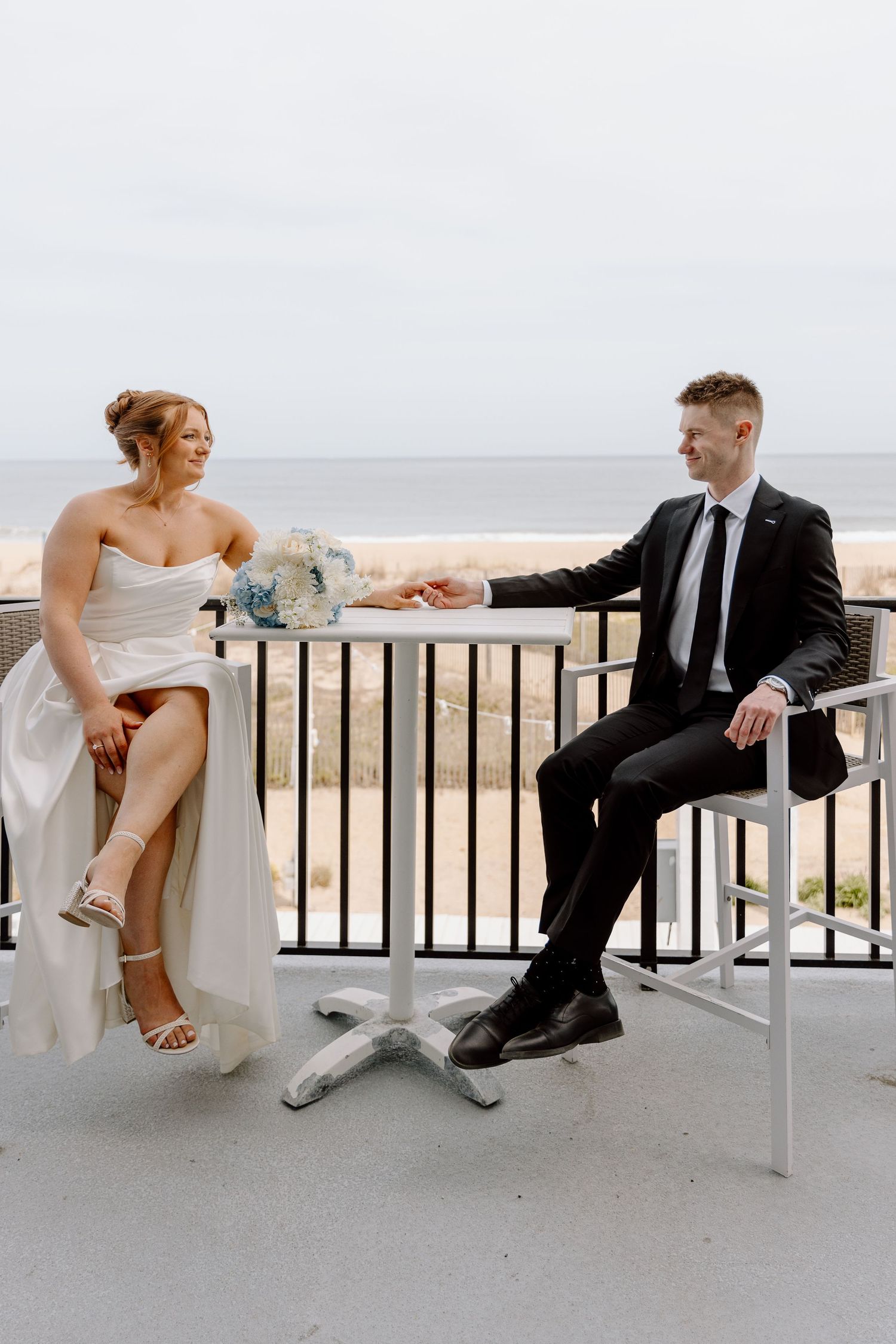 A couple in wedding attire sitting and chatting on outdoor patio chairs by the ocean.