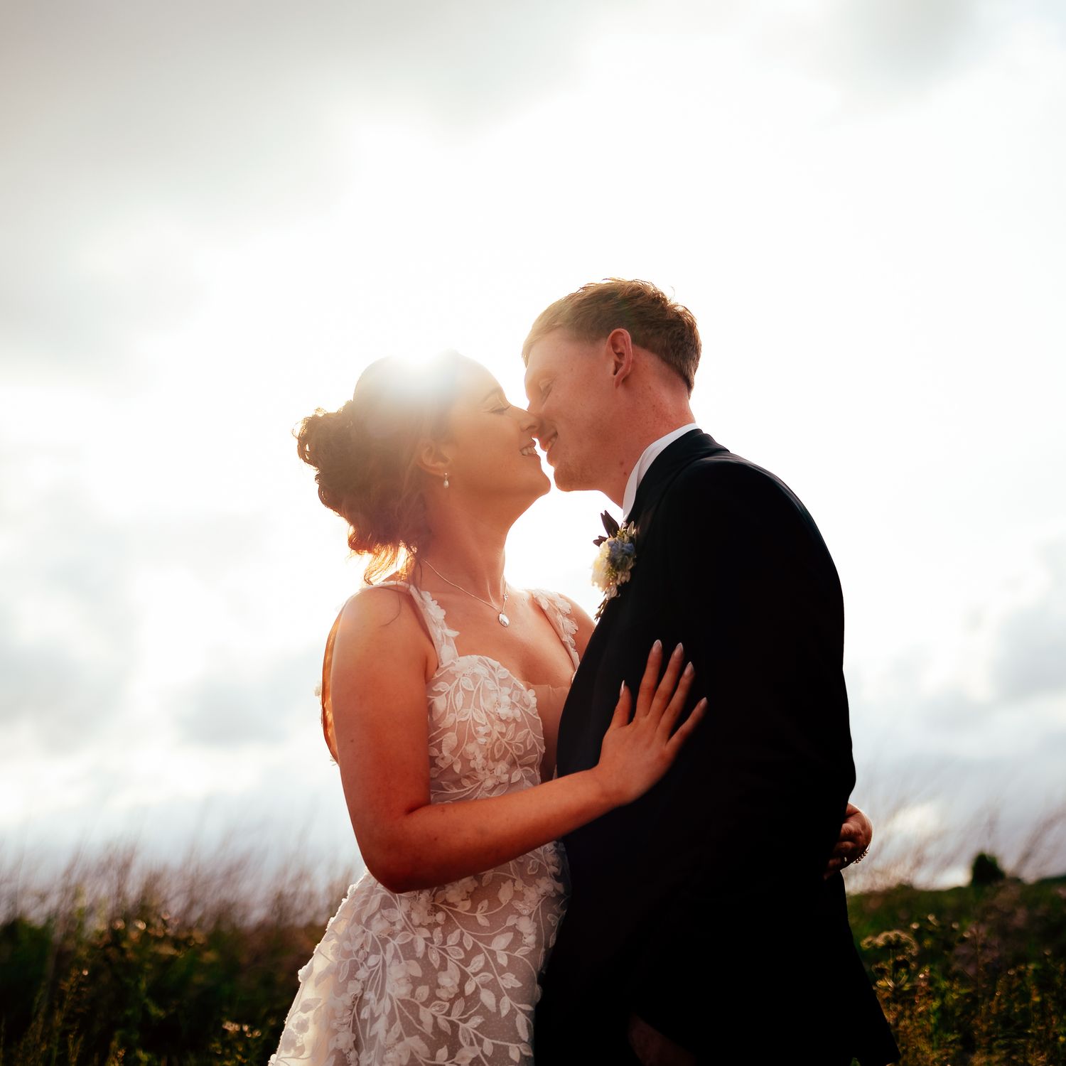 Side profiles of a couple in formal wedding attire kissing at sunset with dramatic cloudy sky in the background.