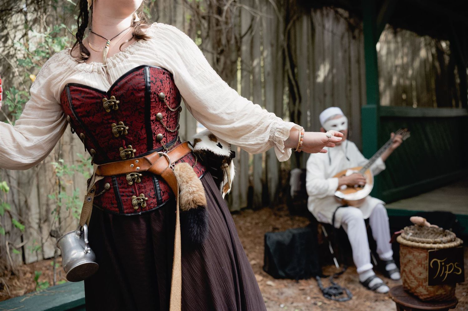 A figure in a Victorian-era steampunk dress and corset pours tea from an ornate teapot in a rustic outdoor setting.