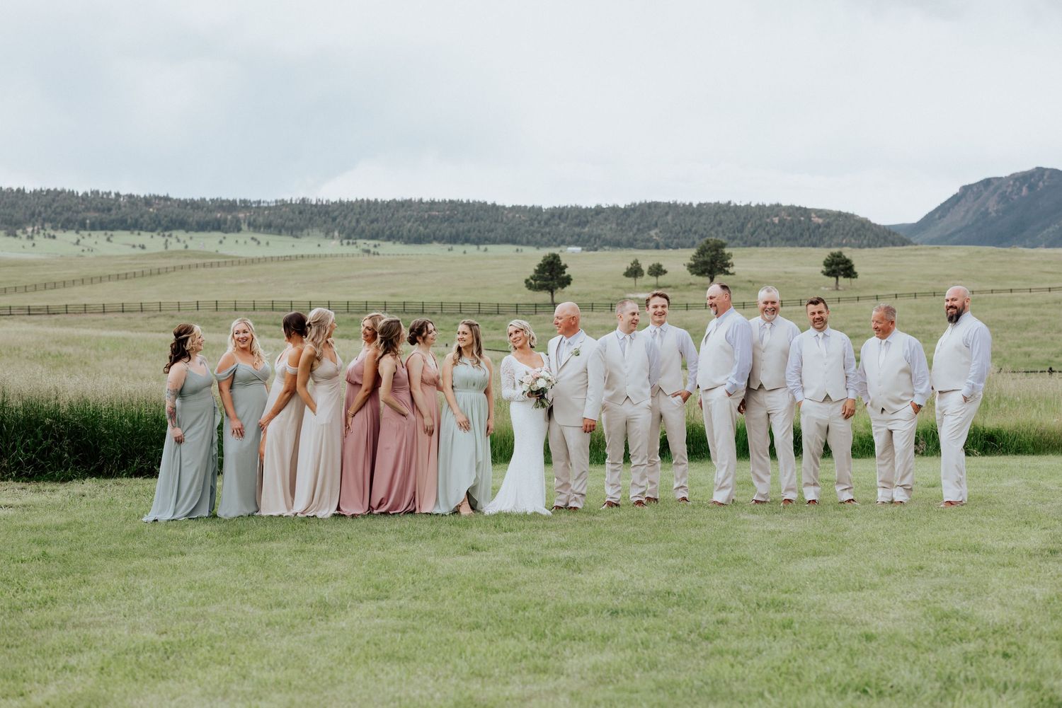 A wedding party in pastel bridesmaid dresses and light colored suits poses together on a grassy field with mountains behind them.