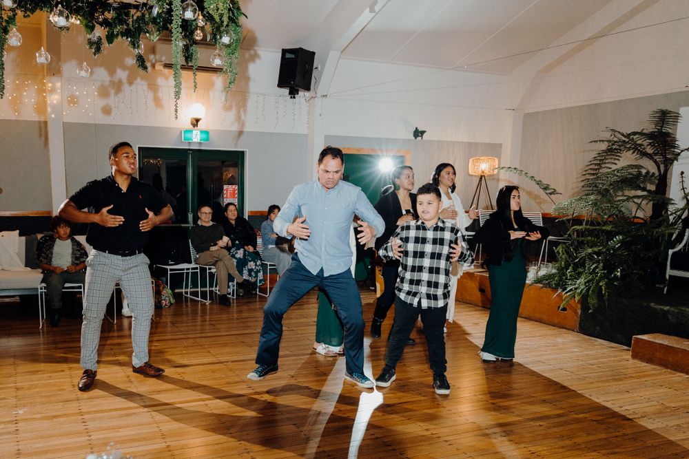 Group dance performance in a bright reception venue with wooden floors and hanging greenery.