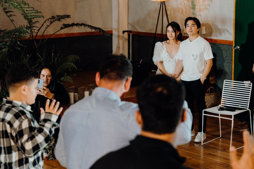 Couple poses in front of indoor plants while group of people watch in casual white and modern setting.