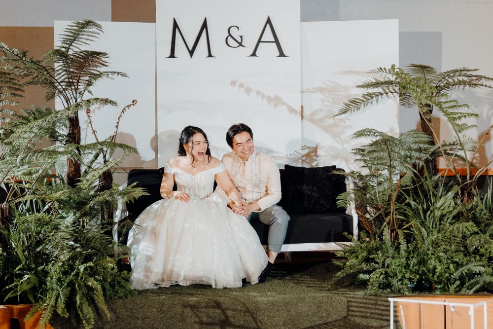 A couple sits on a black piano bench at their wedding with M&A signage and ferns in the background.