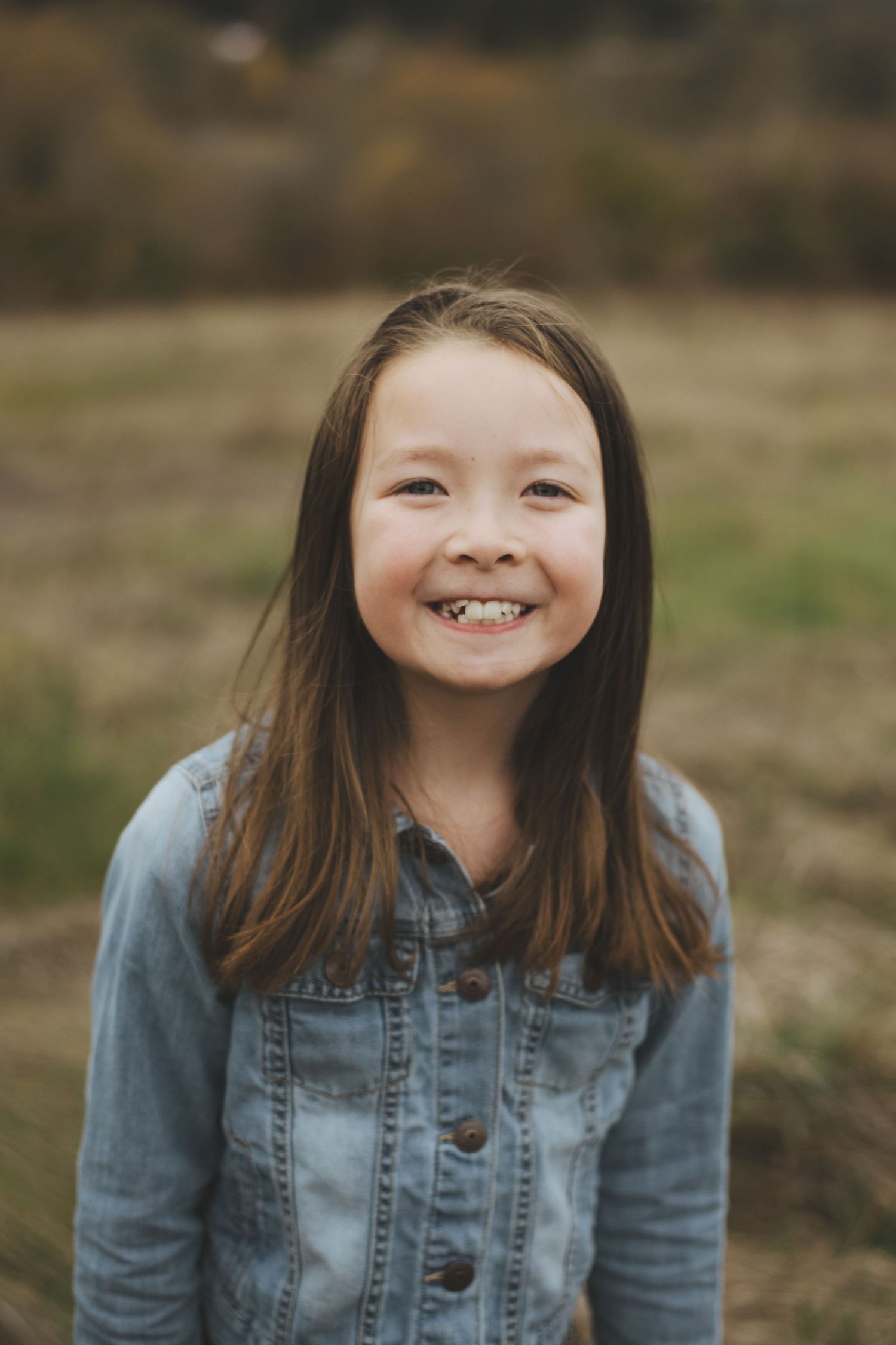 A young child wearing a denim jacket smiles brightly outdoors against a blurred natural background.