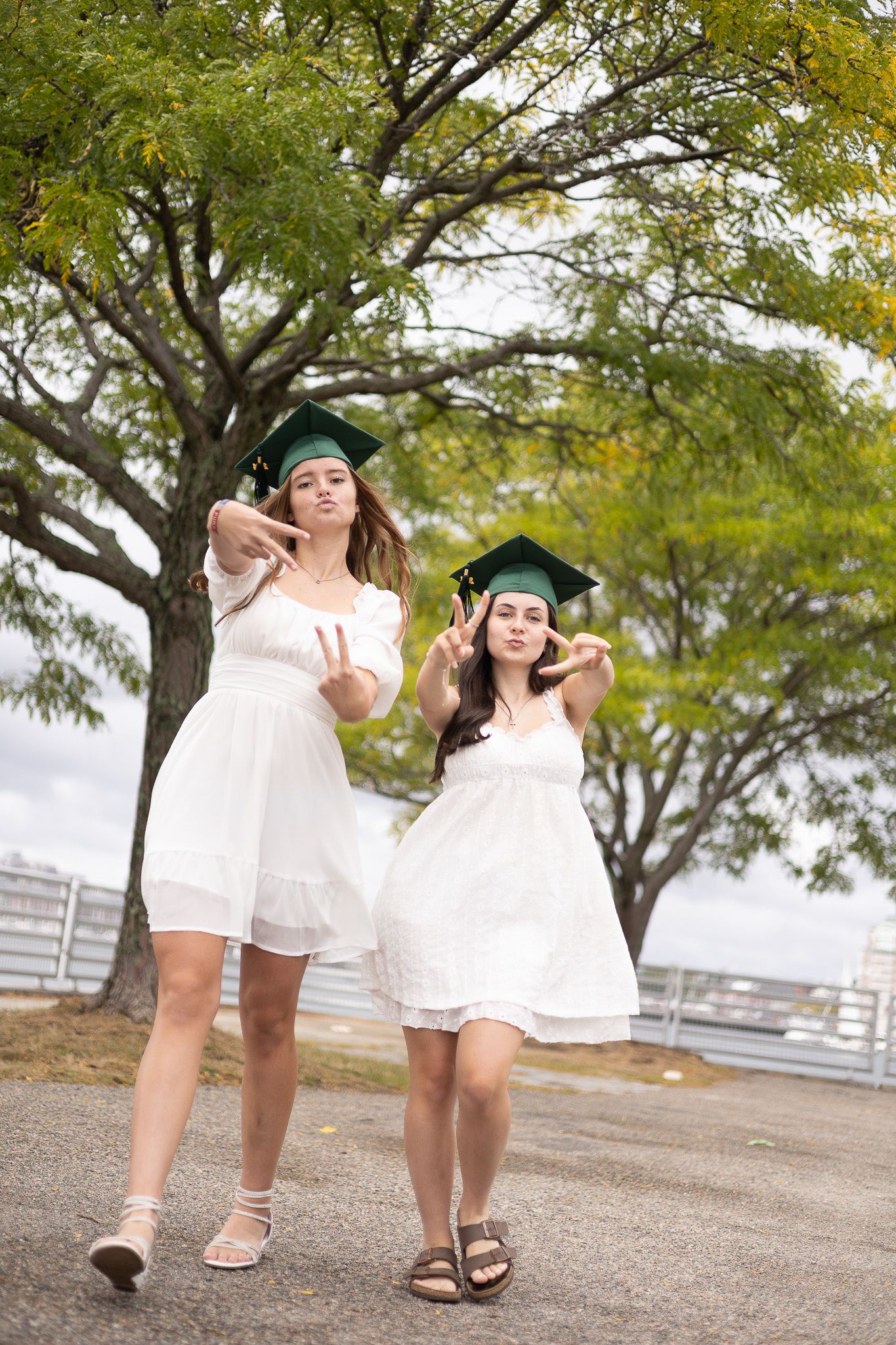Two graduates in white dresses and graduation caps pose playfully outdoors under trees on their celebration day.