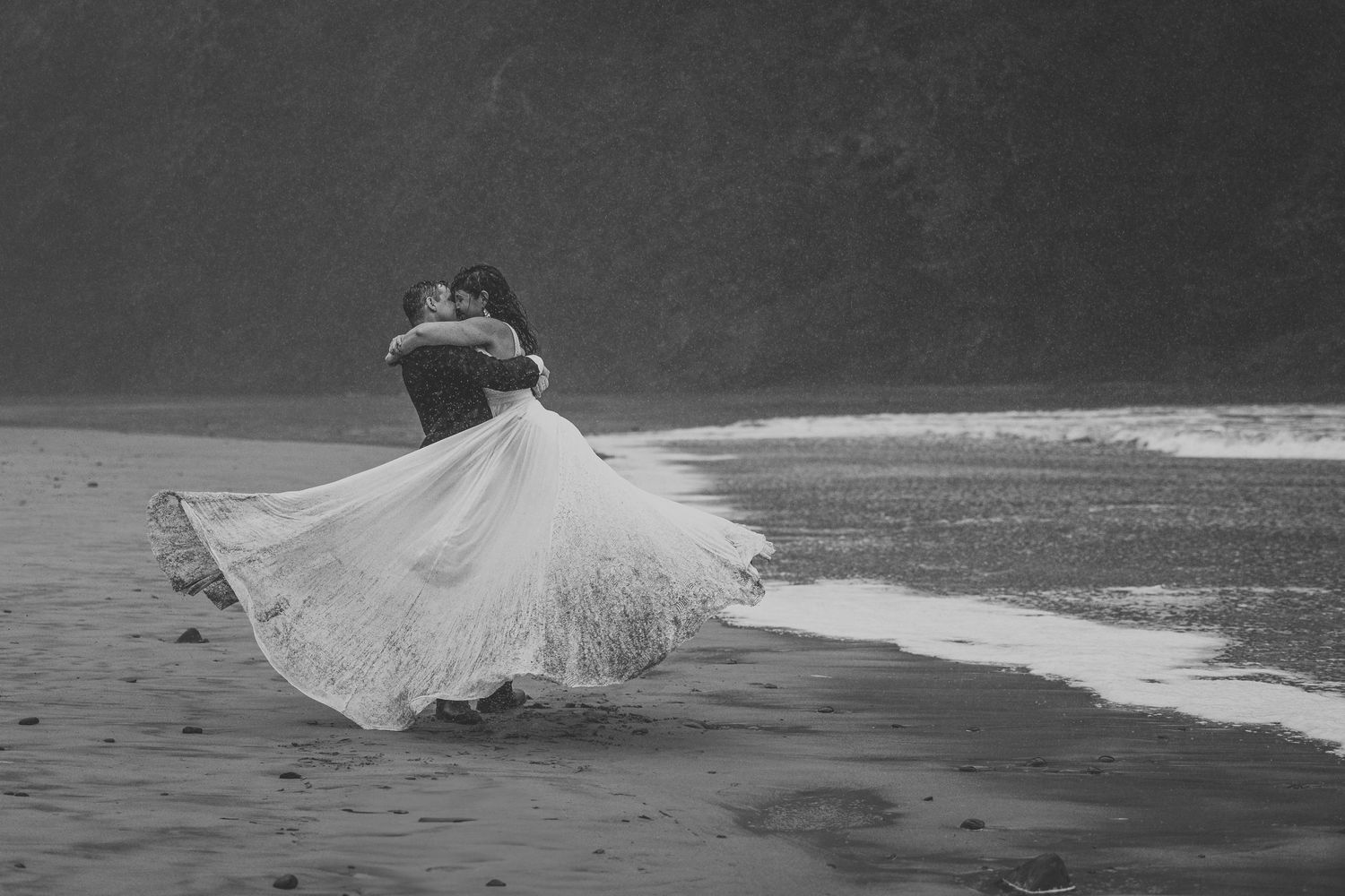 A couple dances on a misty beach as the bride's flowing white dress gracefully twirls in the wind.