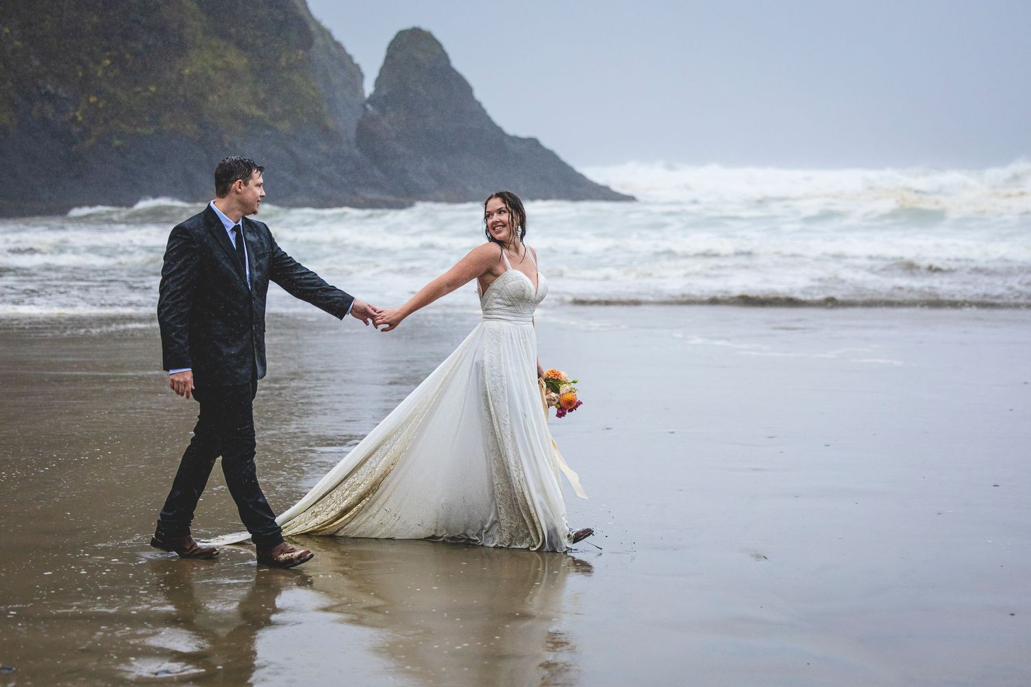 Newlywed couple walks hand in hand along misty beach shore with dramatic coastal cliffs in background.