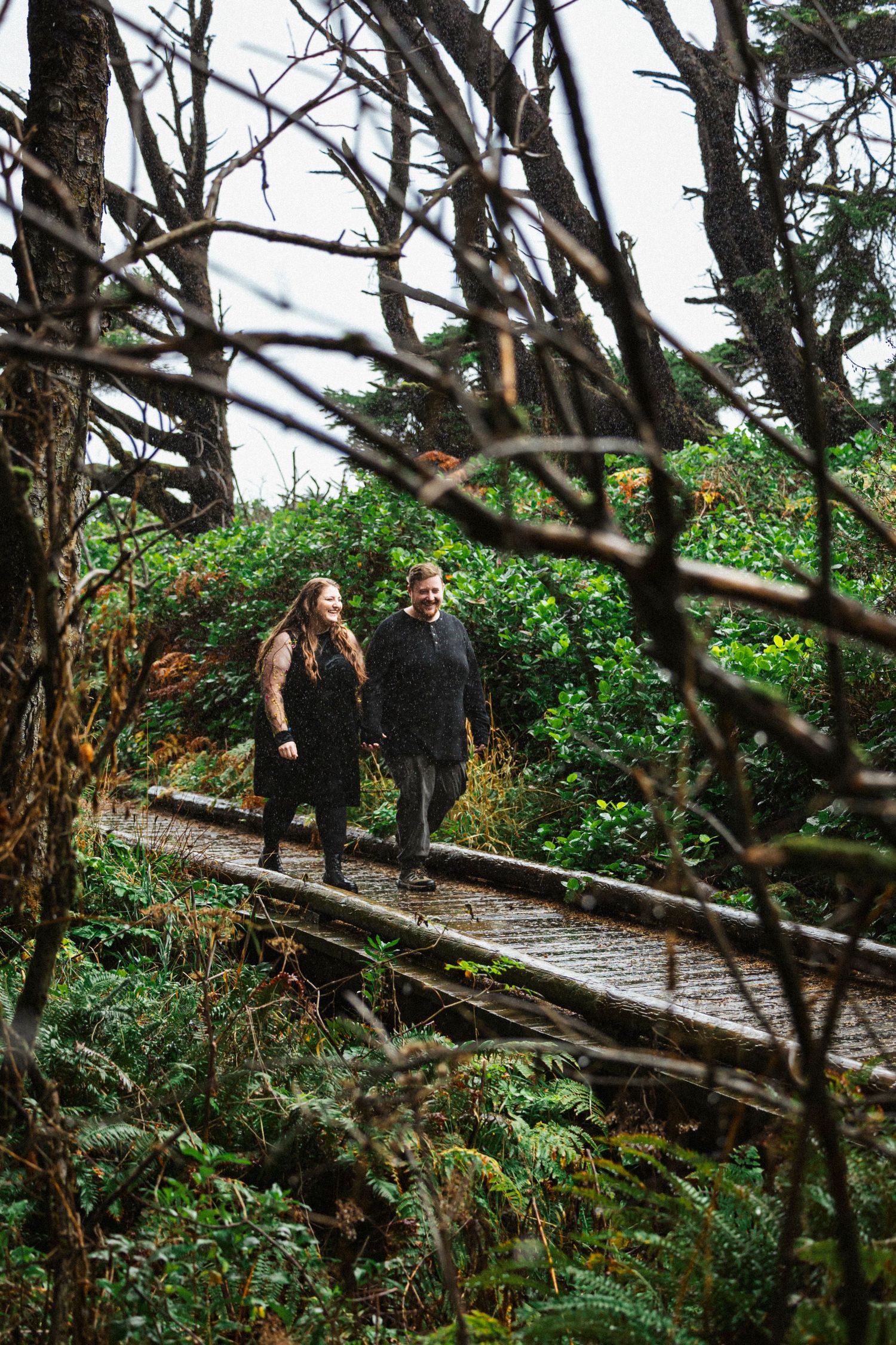 Two people walking together on a wooden boardwalk trail through lush green forest in Pacific Northwest.