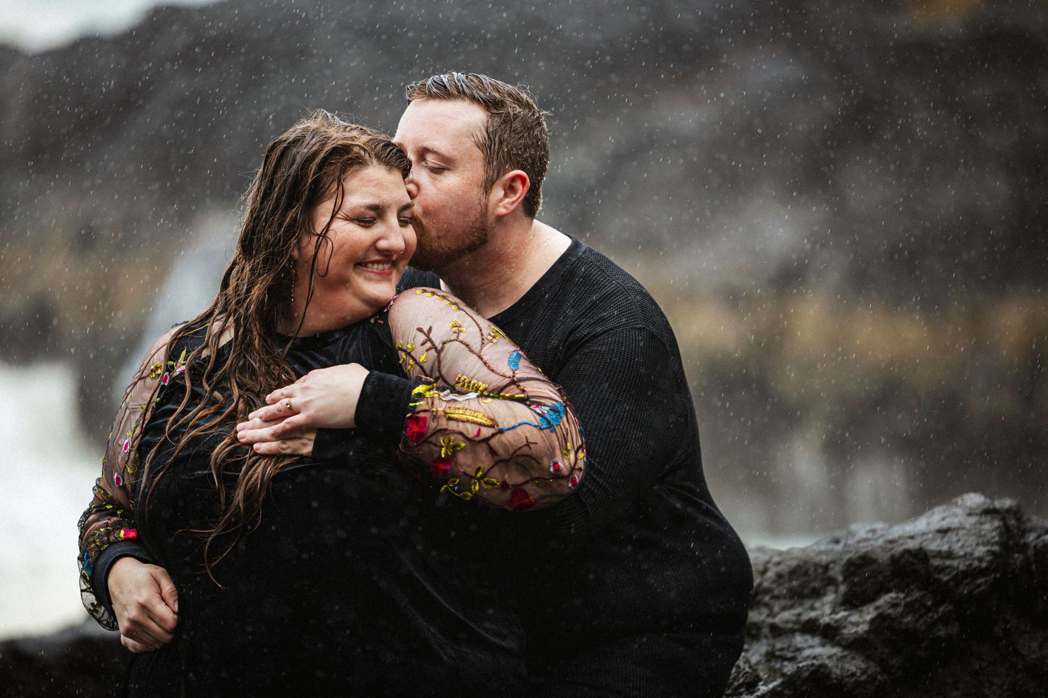 A couple embraces intimately on dark rocks during a moody outdoor photo session in nature.