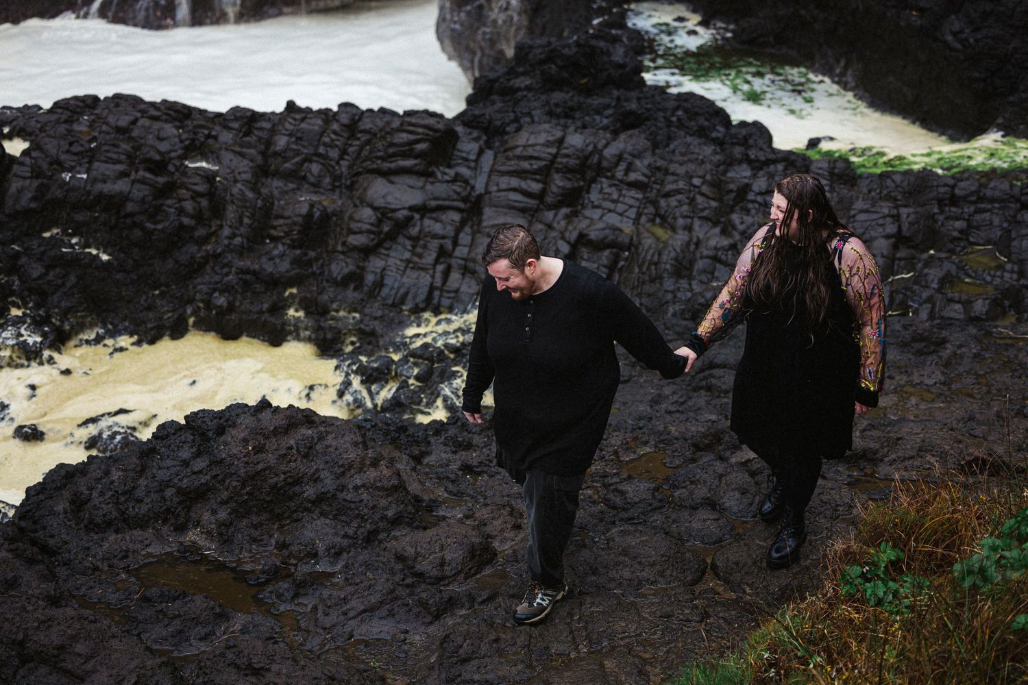 Two people hiking along dark rocky terrain near rushing water and snow patches.