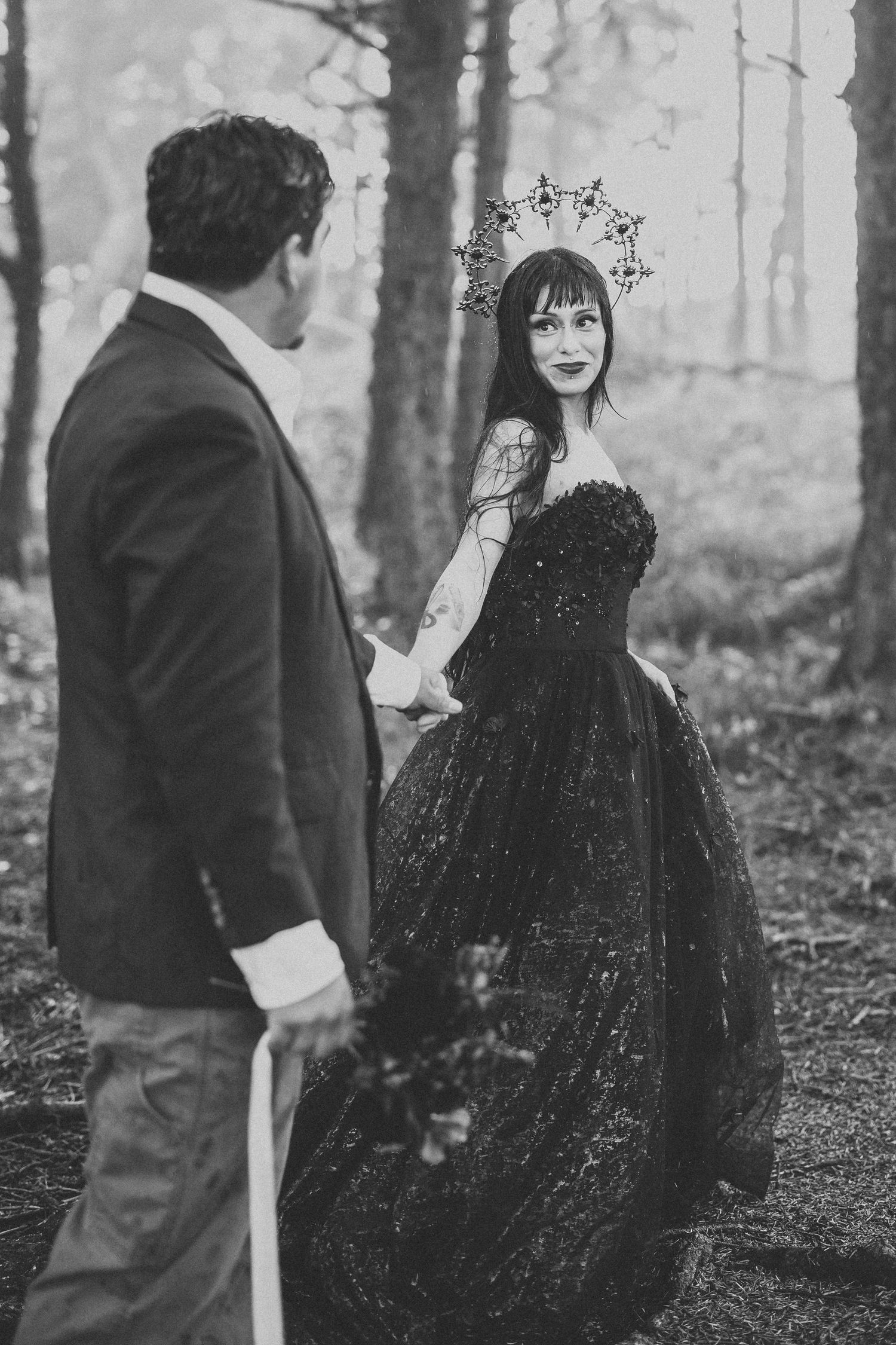 Artistic black and white wedding photo featuring a couple in a misty forest with the bride wearing a gothic black gown.