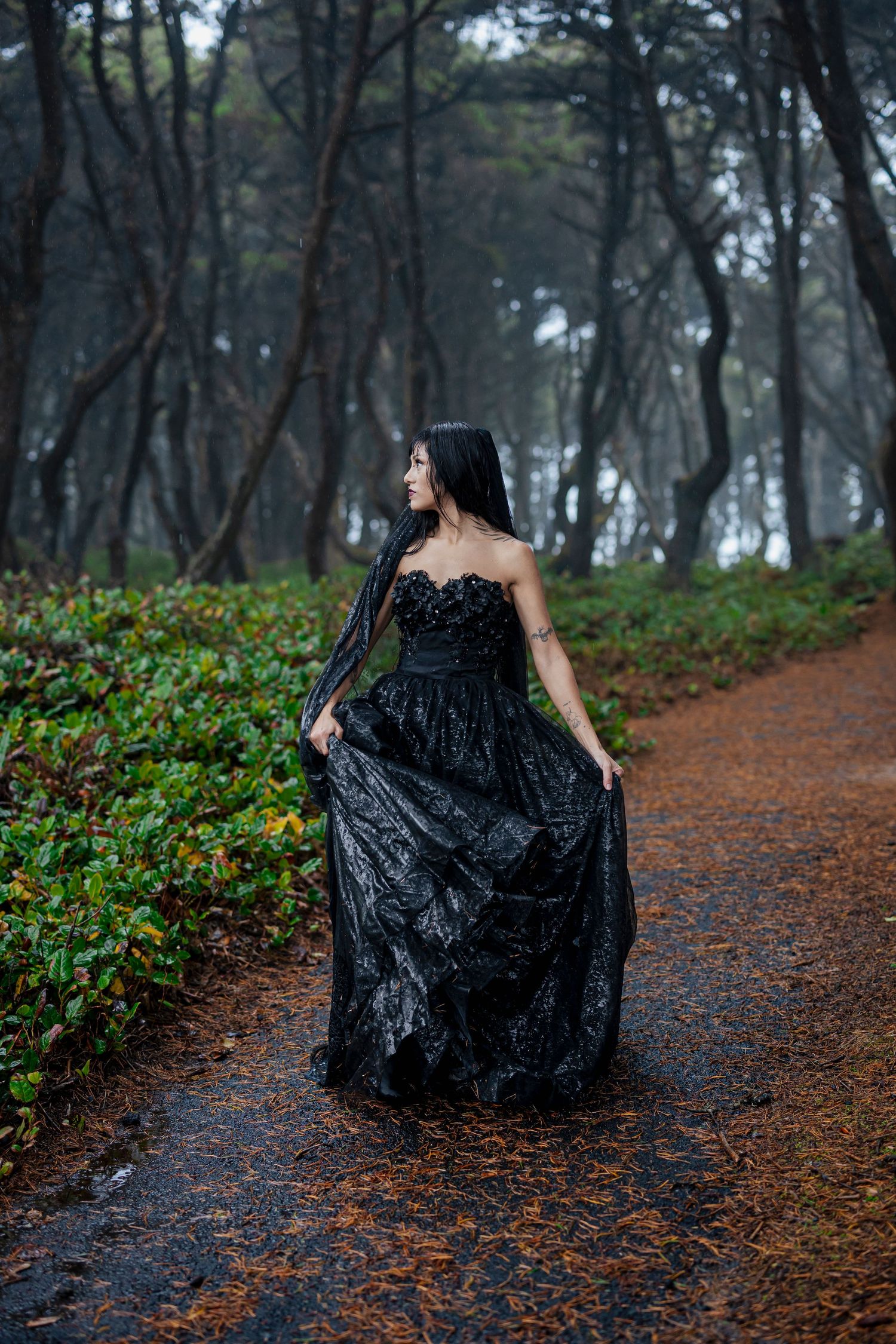 A figure in an elegant black gown stands on a misty forest path surrounded by tall trees and greenery.