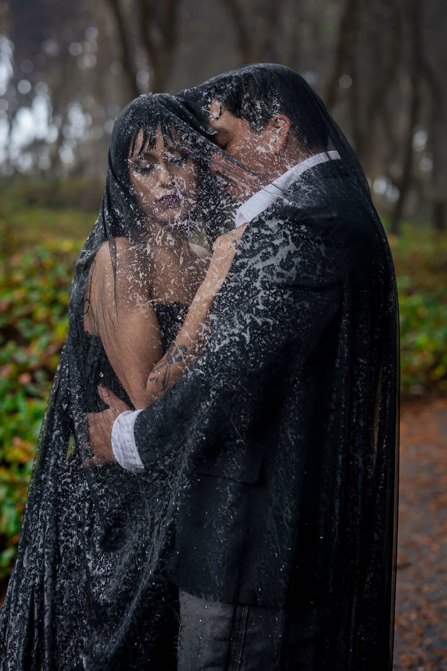 A couple embraces underneath wet black lace in a misty forest setting, creating a moody romantic atmosphere.