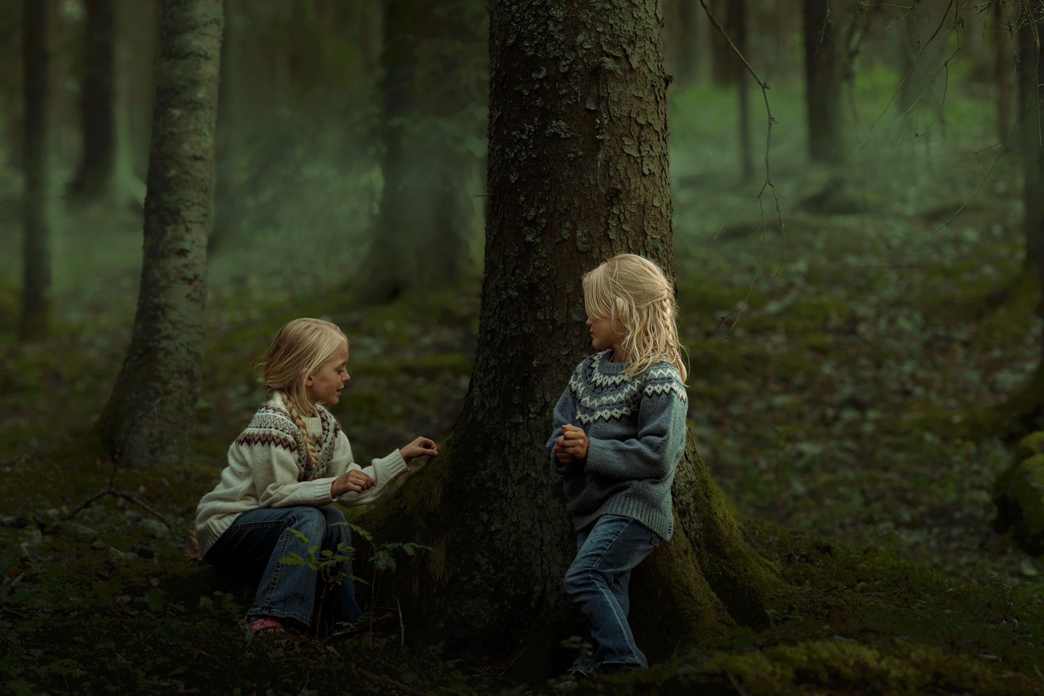 Två flickor på varsin sida om ett träd. De pratar om något. Skogen ser förtrollad ut.