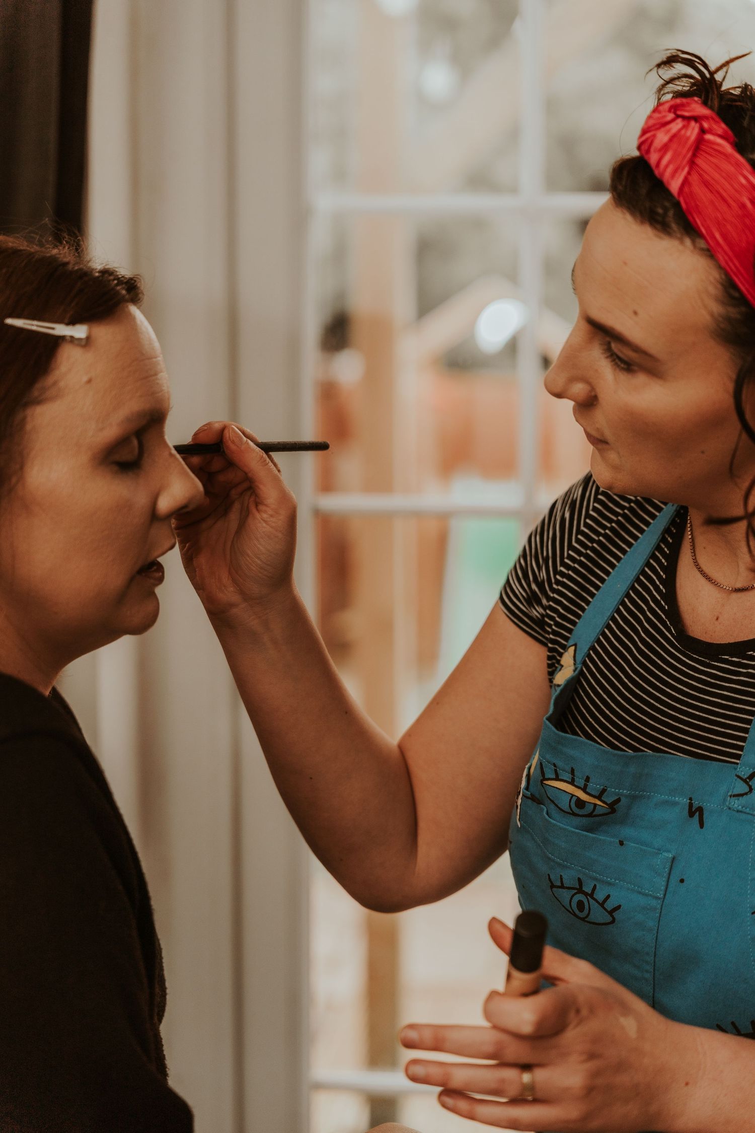 Makeup artist wearing blue overalls and red headband applies eye makeup with brush in warm indoor lighting.