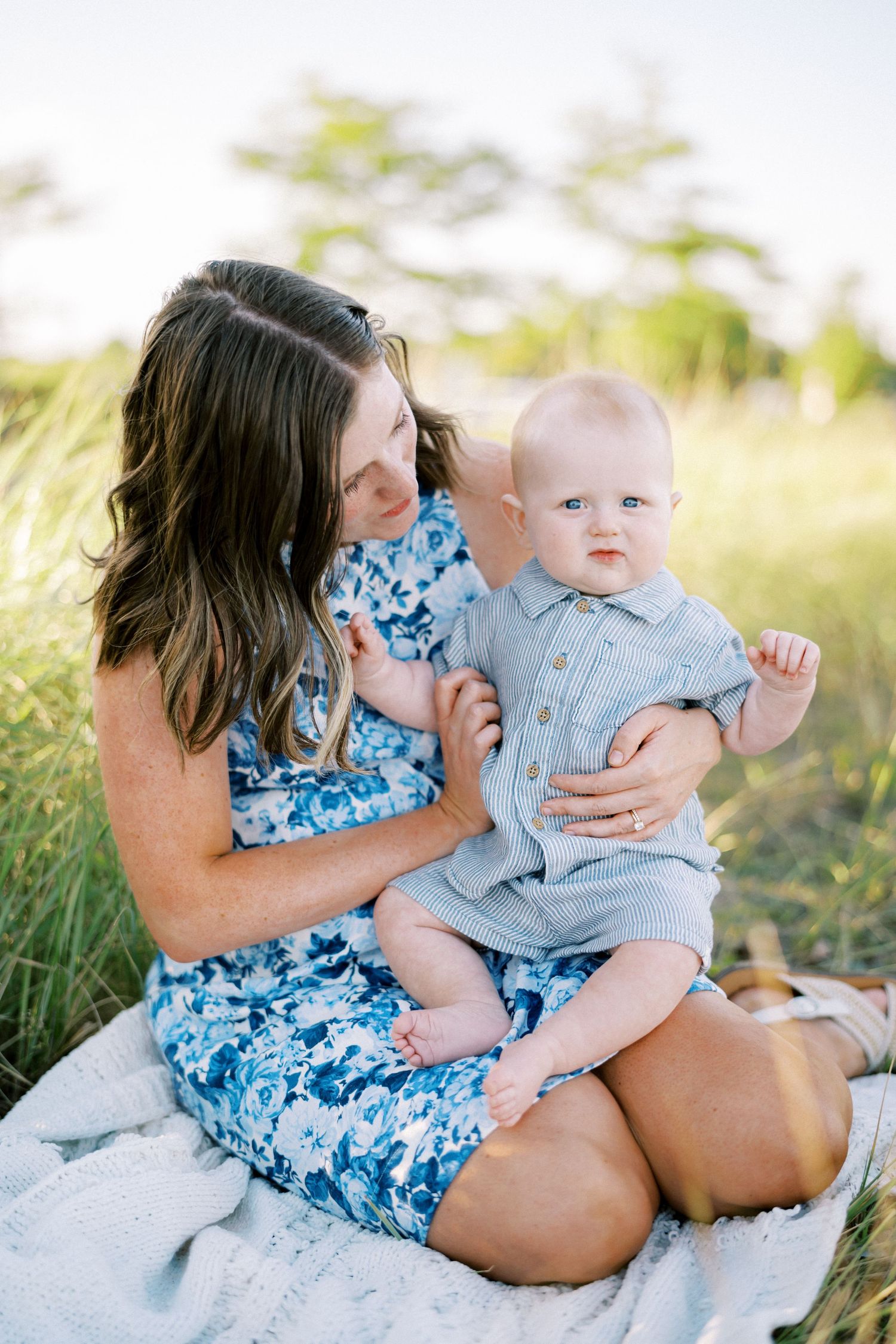 A baby and woman wearing blue sit together on a blanket in a sunny field during a summer photo session.