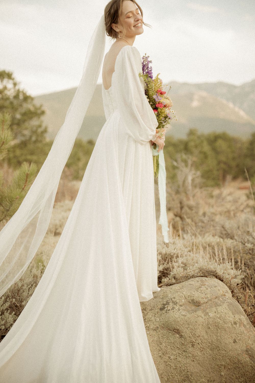 Elegant bridal portrait of a white wedding dress with long flowing veil and colorful bouquet against mountain backdrop.