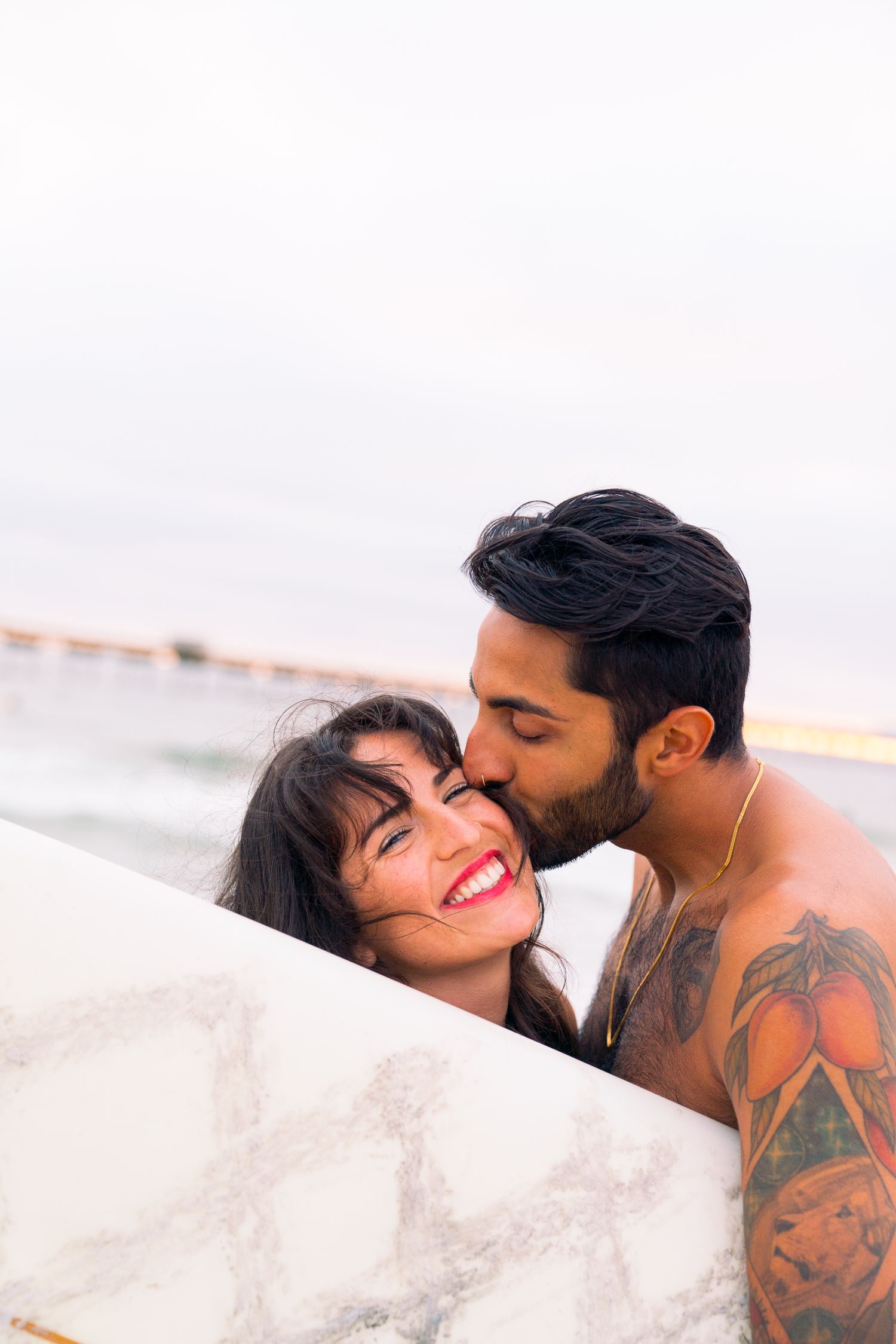 A romantic couple shares an intimate moment behind a white wall near a beach at sunset.