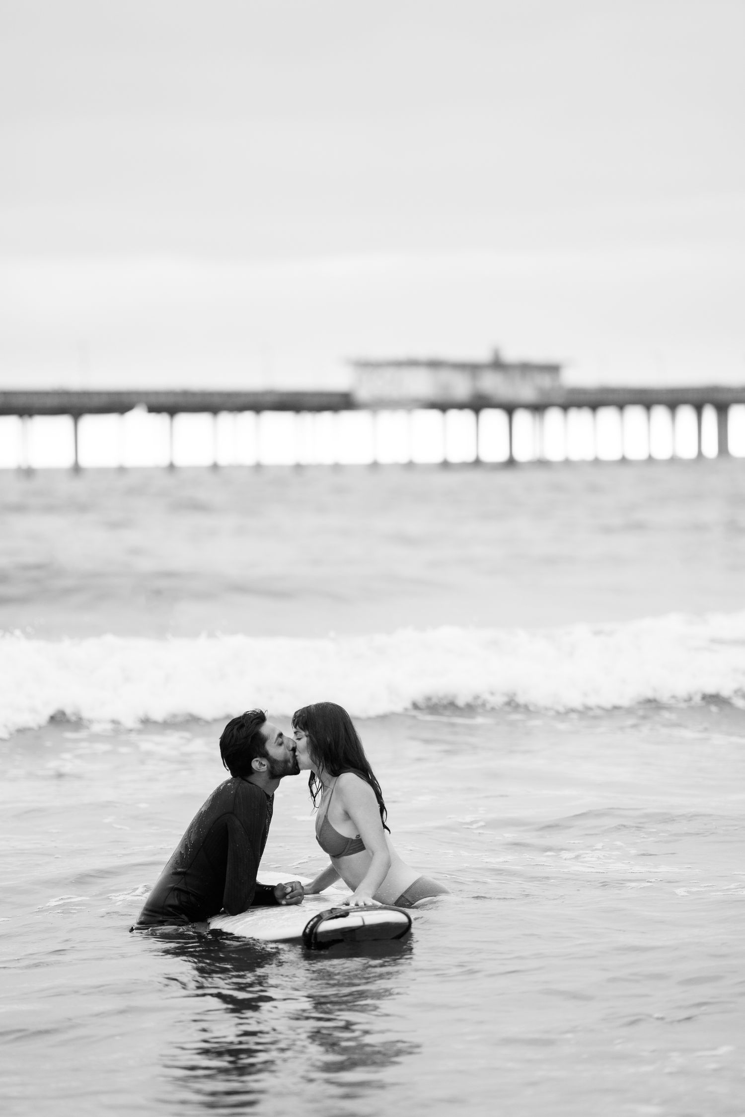 A couple shares a romantic moment on surfboards near a pier in black and white.